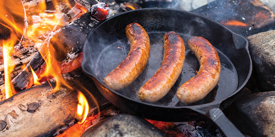 Sausages cooking in cast iron skillet over campfire